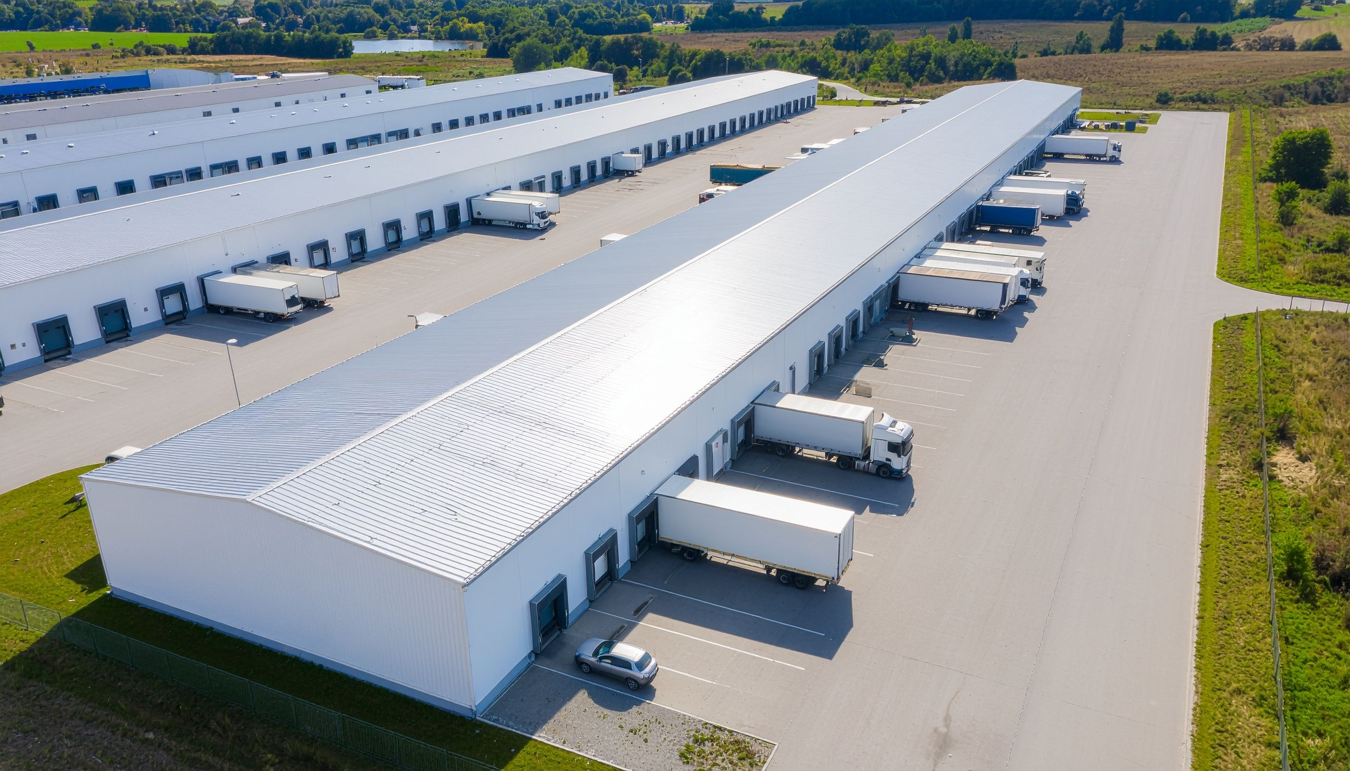 Luftaufnahme eines großen Industrie- und Logistikkomplexes mit langen weißen Hallenbauwerken, Laderampen und mehreren LKW auf dem Betriebsgelände