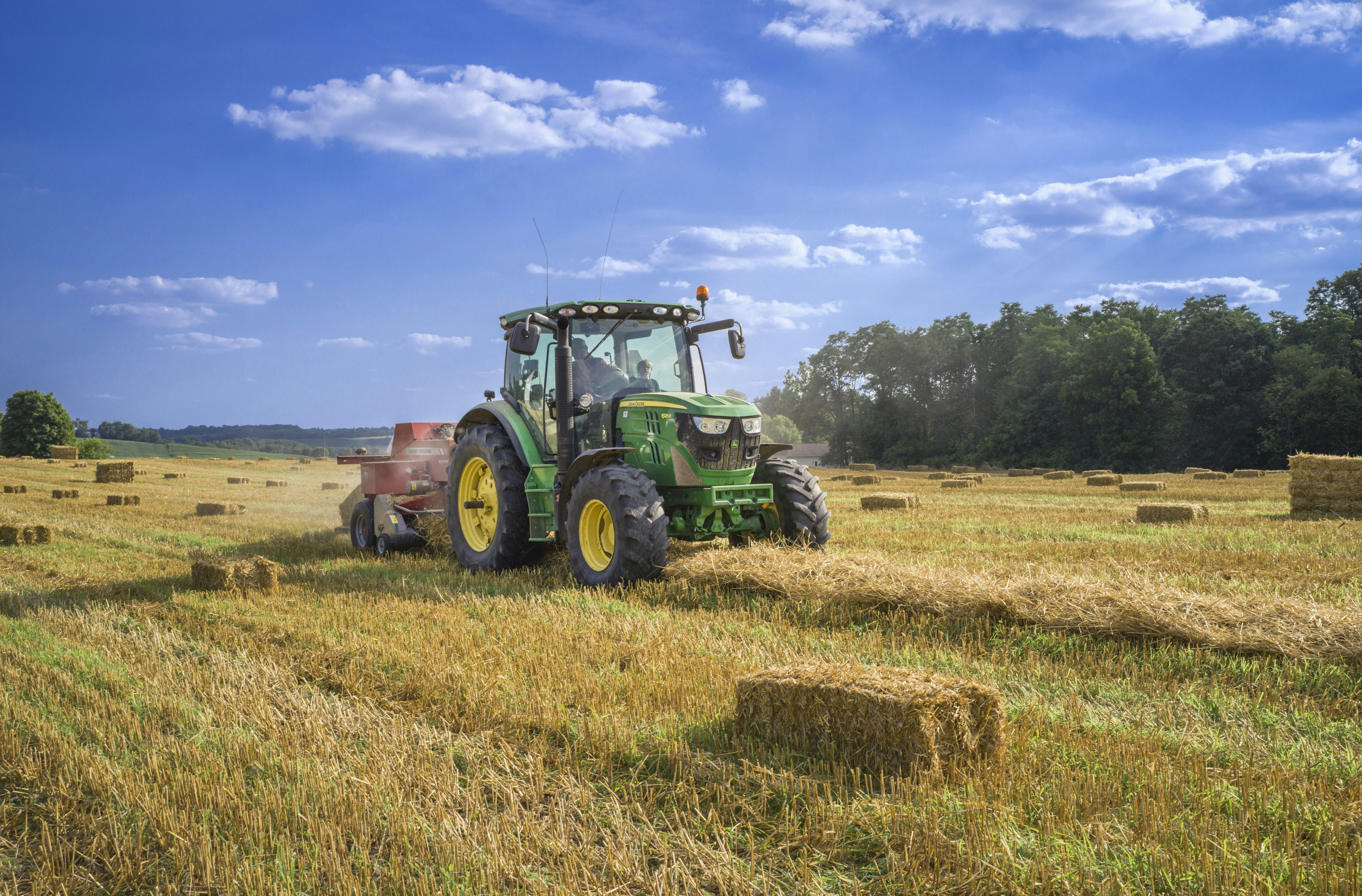 Grüner Traktor fährt über ein abgeerntetes Feld mit Strohballen unter blauem Sommerhimmel