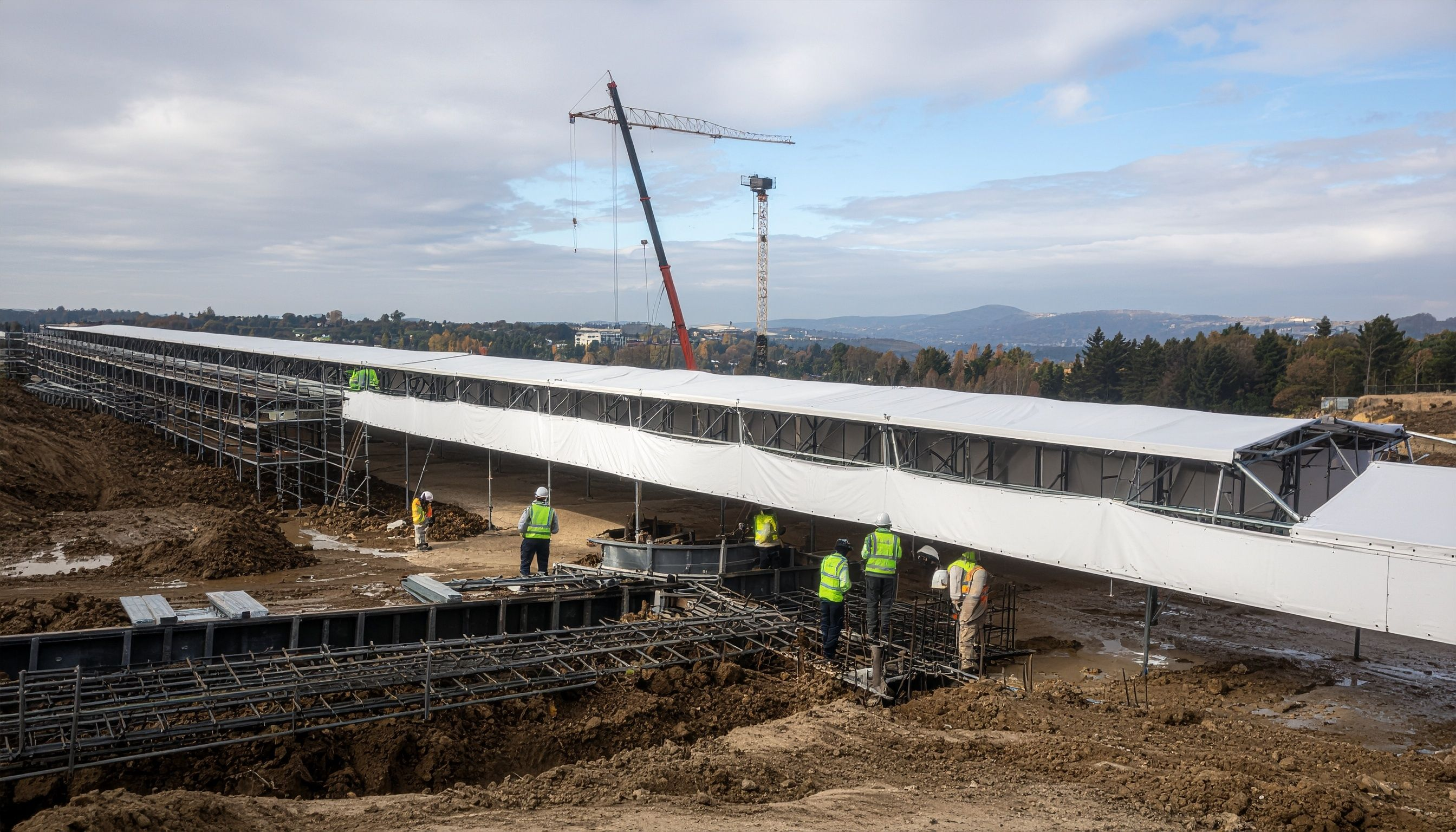 Langes weißes Baustellenzelt auf einem aktiven Bauprojekt mit Bauarbeitern in Warnwesten, Fundamentarbeiten im Vordergrund und Baukran im Hintergrund